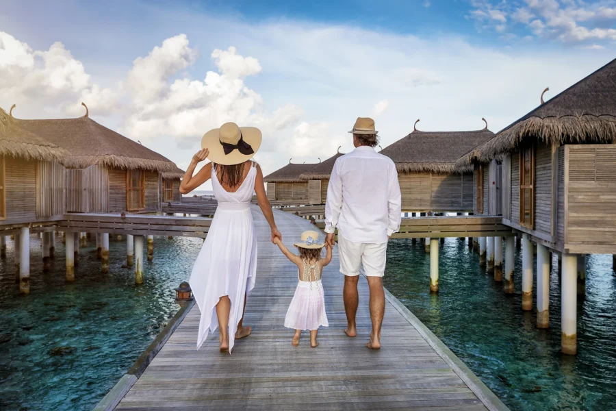A family in white summer clothes walks over a wooden pier between over water lodges in the Maldives